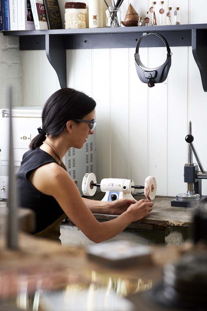 A Woman sitting at a tool bench, using a polishing motor.